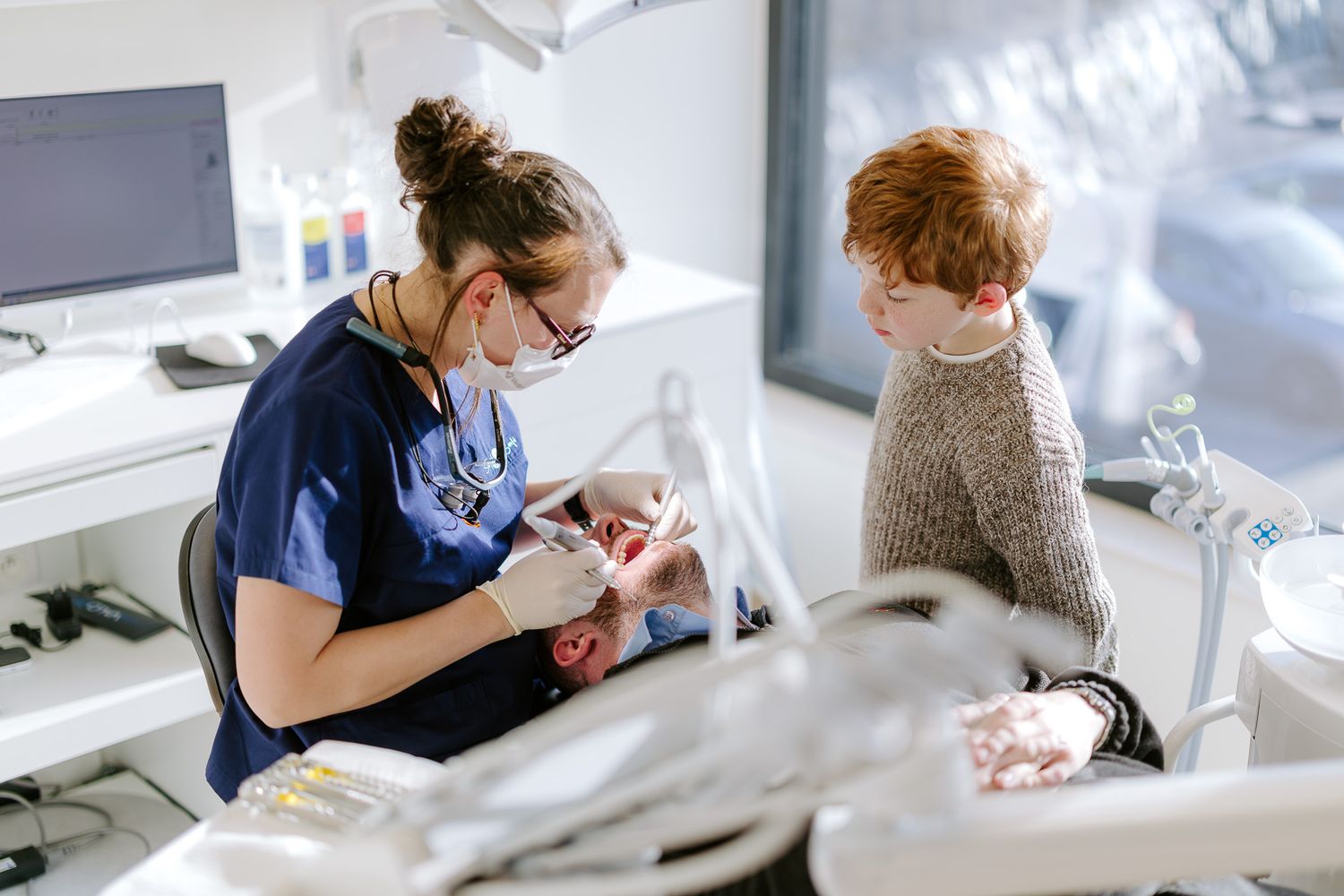 Female dentist in mask shows dental model to young red-haired child patient in dental clinic.