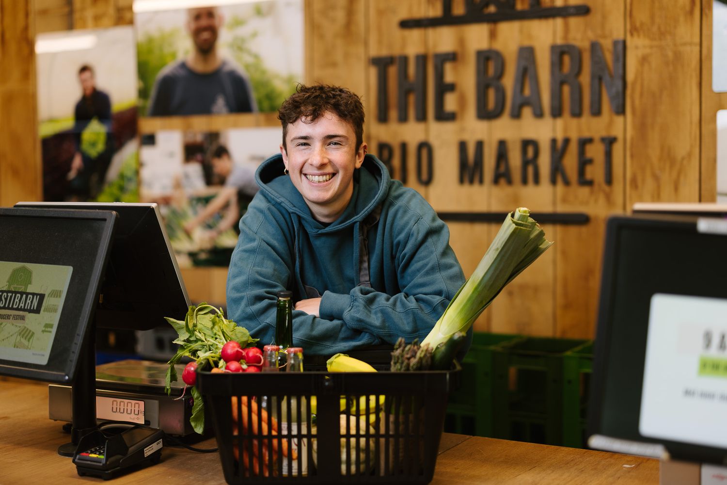 Smiling young man at checkout of The Barn Bio Market with a basket of fresh vegetables.