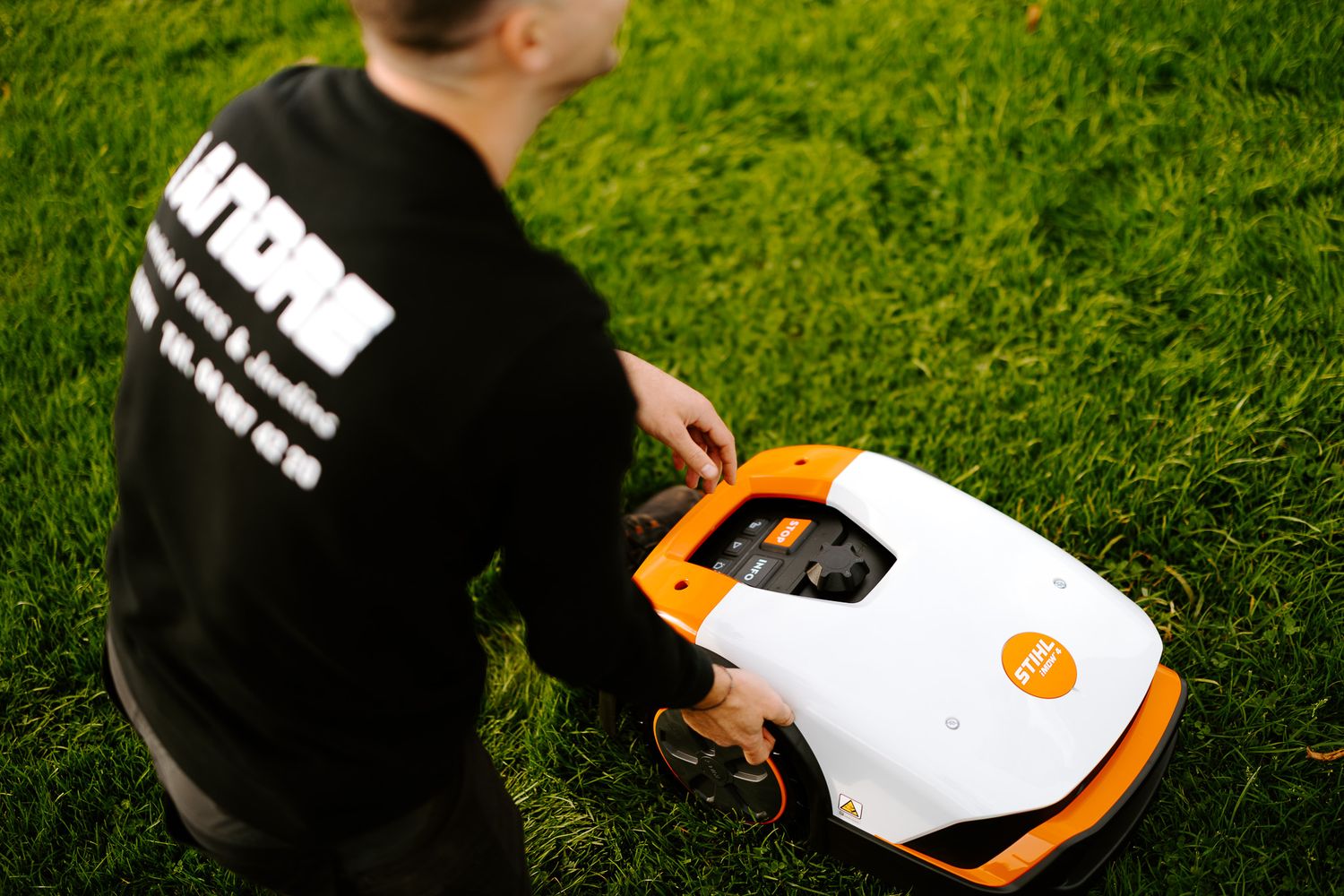 Technician in black uniform kneeling on grass while servicing an orange robotic lawn mower.