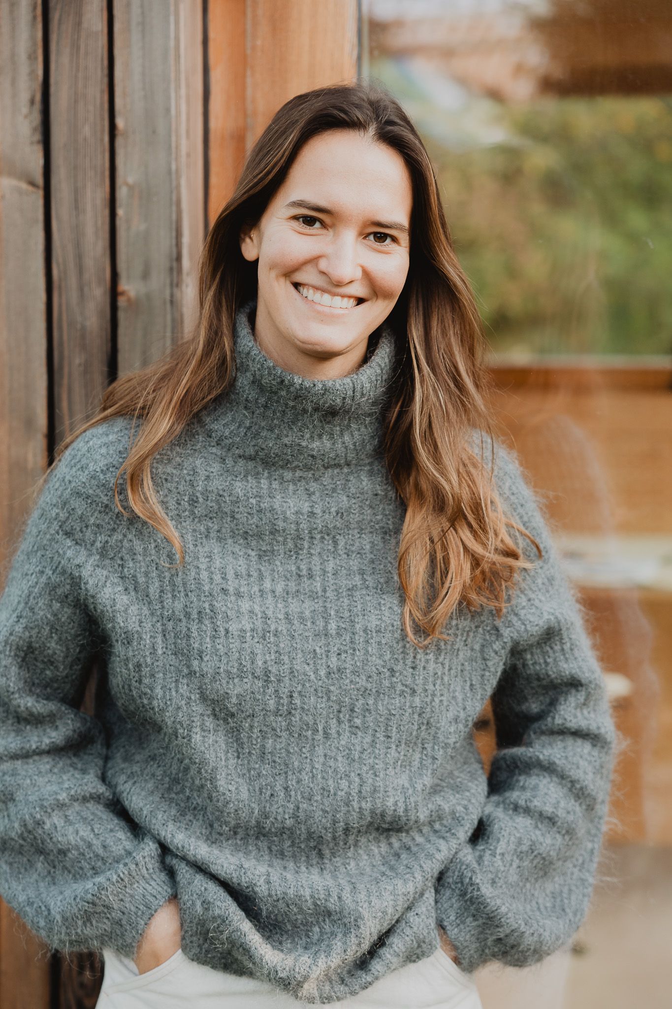 Smiling woman with long brown hair wearing a cozy gray turtleneck sweater outdoors.