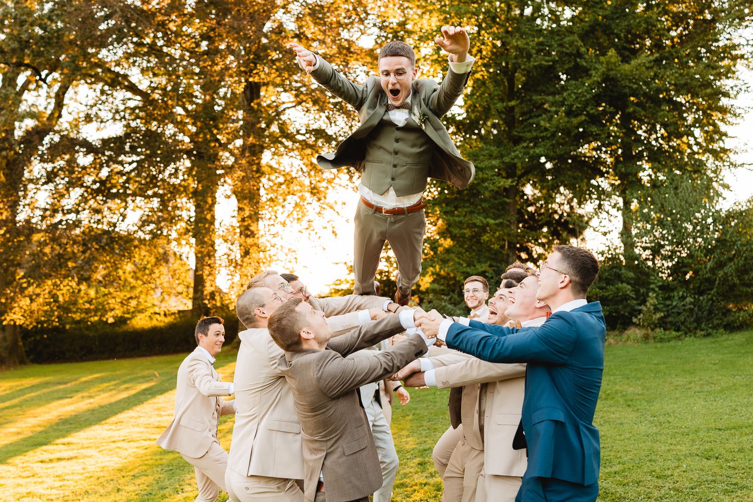 Groomsmen tossing groom into the air in a sunny park during a fun outdoor wedding celebration.
