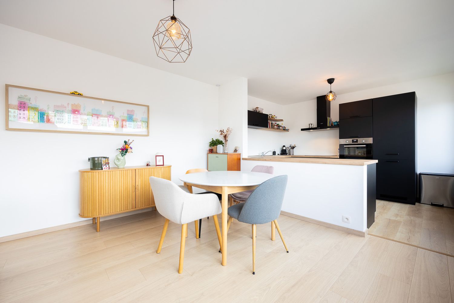 Scandinavian-style dining area with round table, mixed chairs, pendant light, and open kitchen.