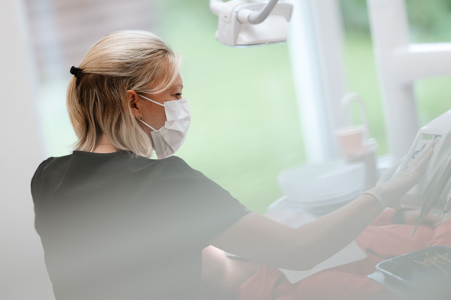 Blonde female dental patient wearing a mask and black cape sitting in a dental chair by a bright window.