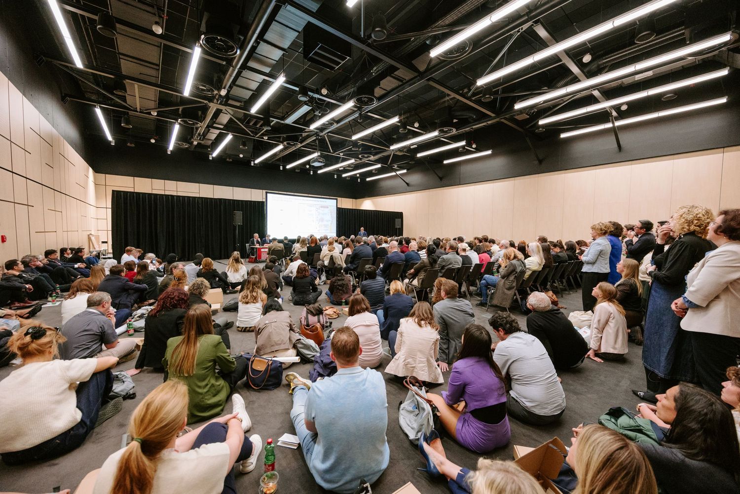 Large audience seated on floor and chairs in modern conference hall during a presentation event.