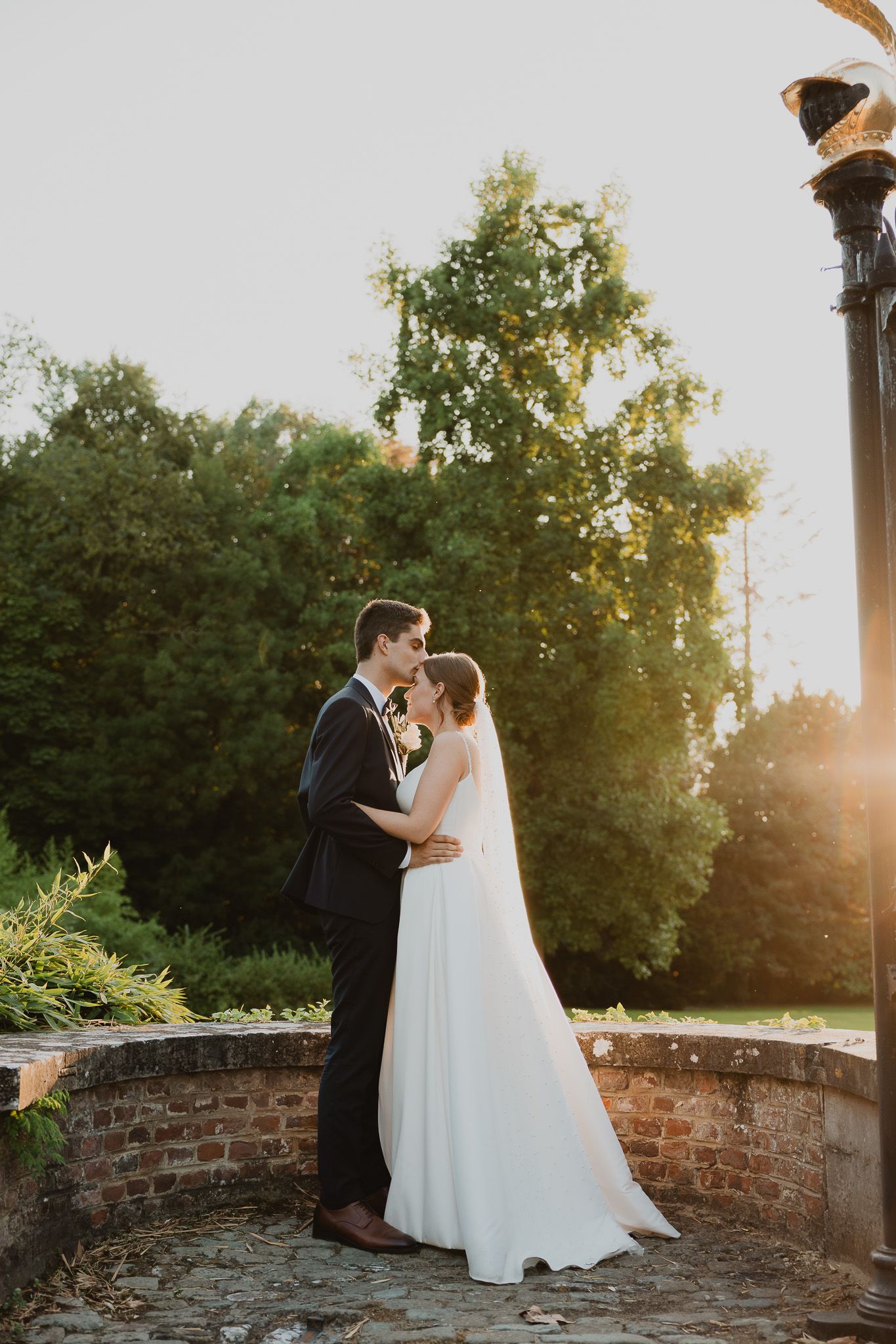 Bride and groom share a romantic kiss at golden hour in a lush garden with brick pathway.