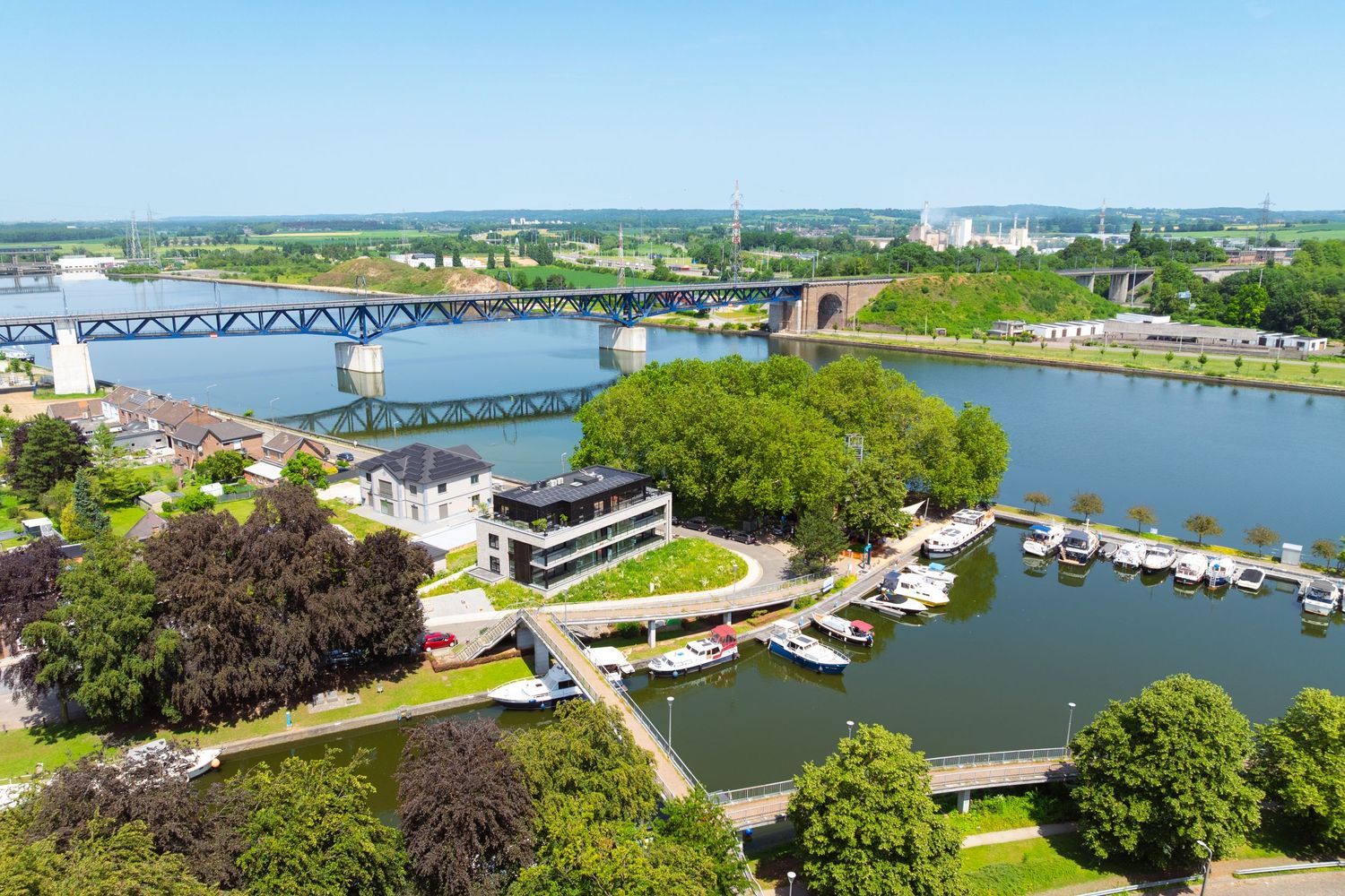 Aerial view of a river marina with boats, green trees, bridges, and industrial buildings under a clear blue sky.