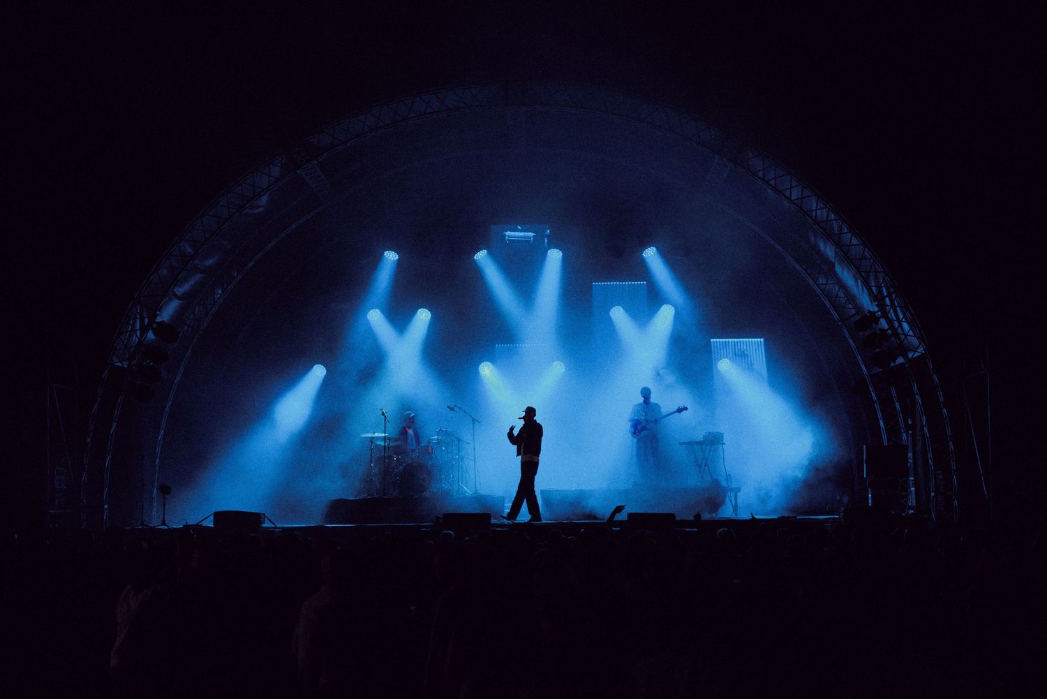 Silhouetted performer on a dramatic blue-lit concert stage with spotlights and band setup under an arched venue.
