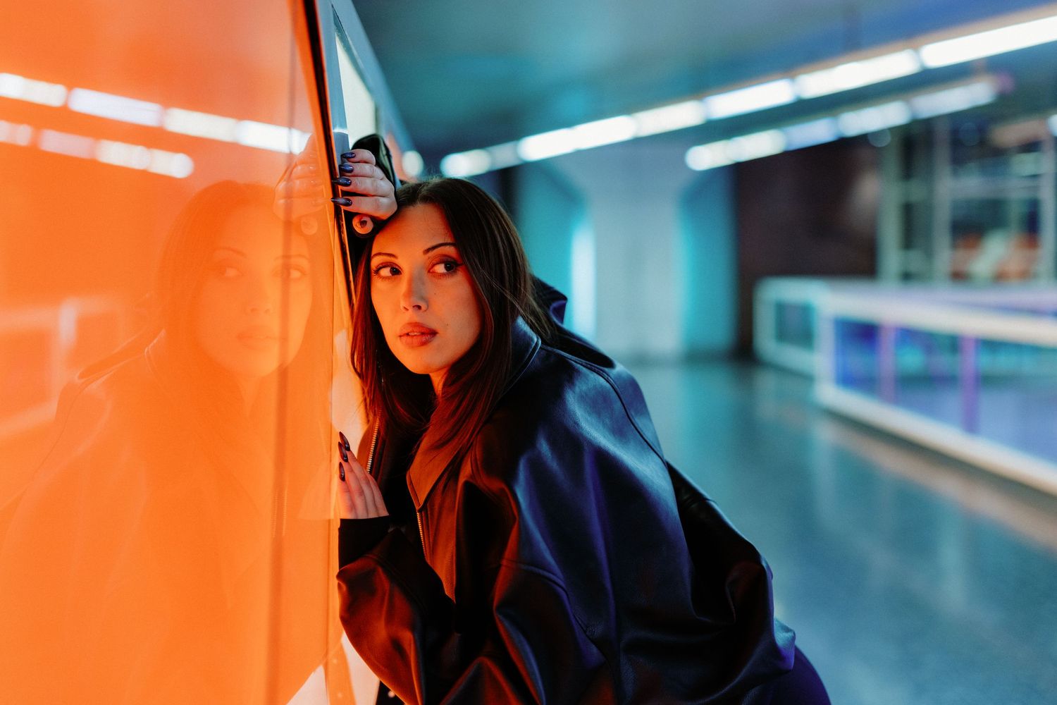 Young woman in dark coat leaning against vibrant orange neon-lit wall in modern underground corridor.
