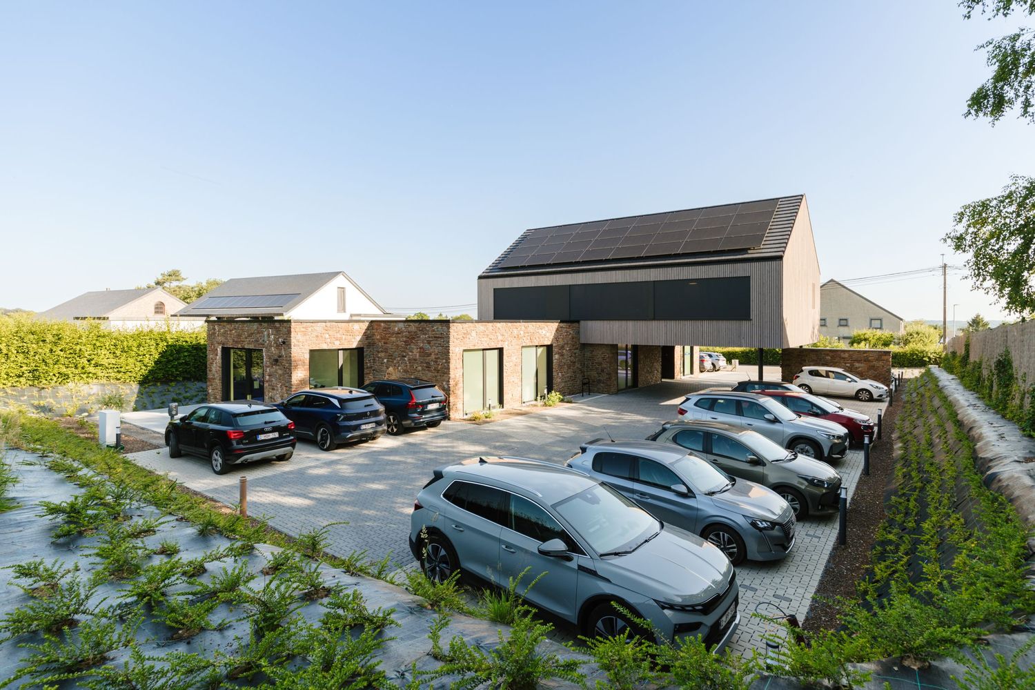 Aerial view of a modern building with a full parking lot surrounded by greenery on a sunny day.