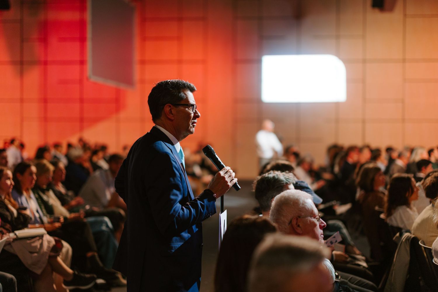A man in a blue suit speaks into a microphone at a large conference, with an audience seated in rows behind him.