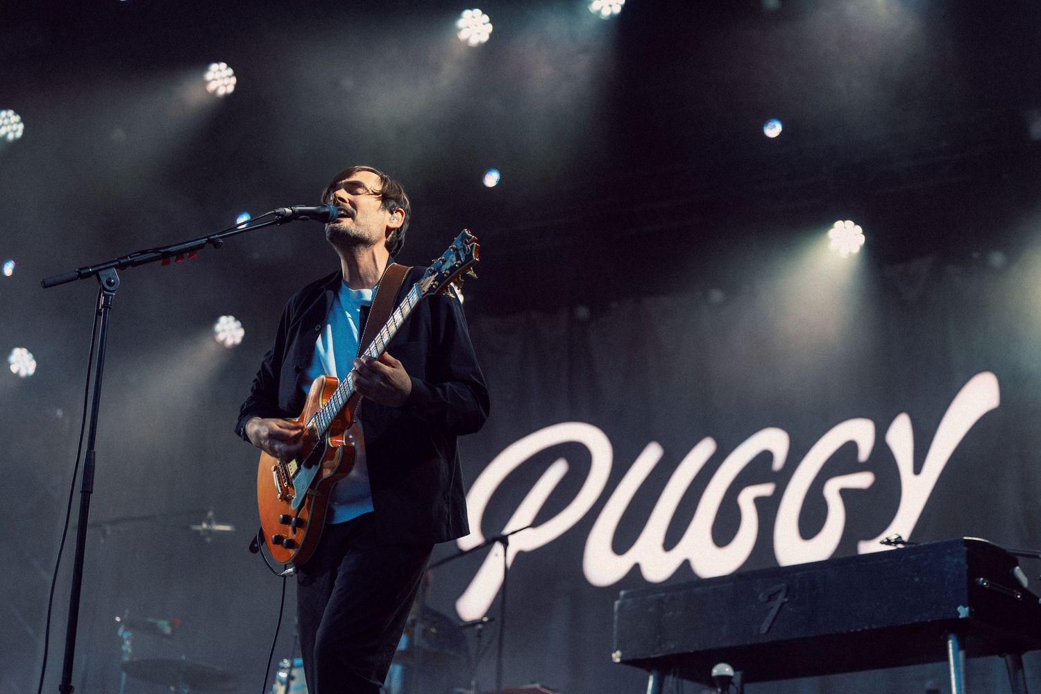 Puggy band guitarist performing live on stage under bright spotlights with band name sign in background.