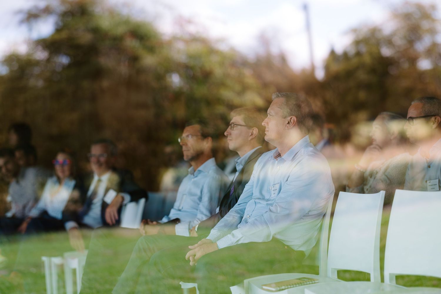 Group of attendees seated outdoors at a corporate event, viewed through a glass reflection.