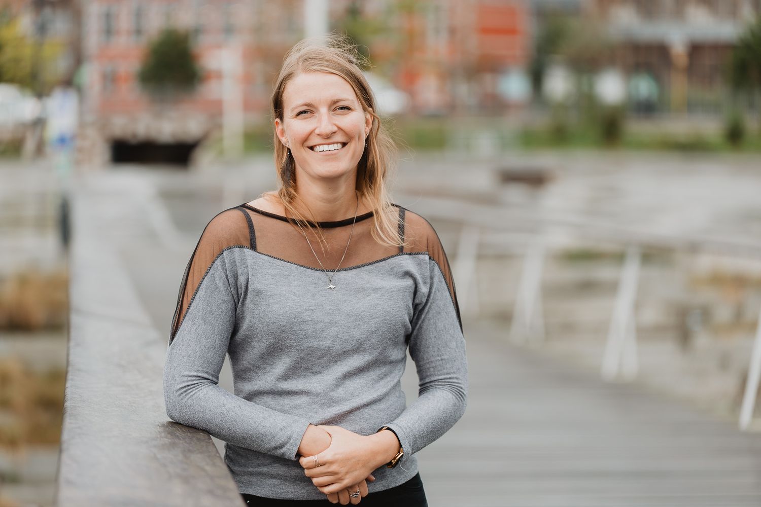 Smiling blonde woman in a gray off-shoulder top leaning on a railing with an urban waterfront background.