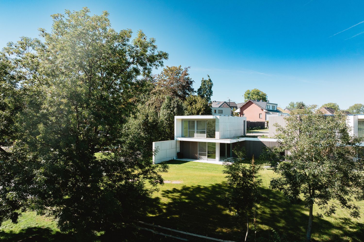 Modern white cubic house surrounded by mature trees and expansive green lawn under blue sky.