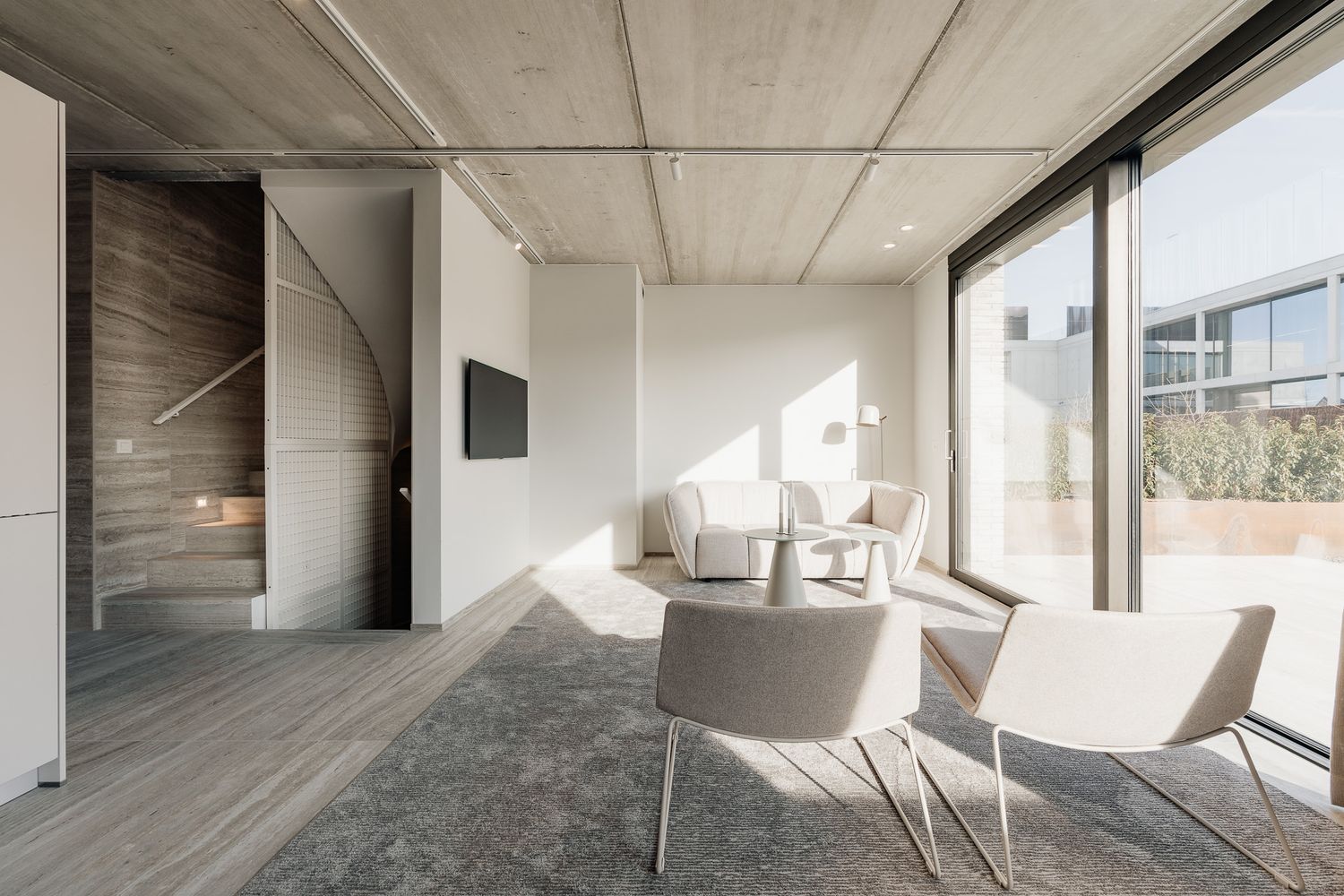 Sunlit modern lounge with concrete ceiling, floor-to-ceiling windows, light chairs, and a wooden staircase.