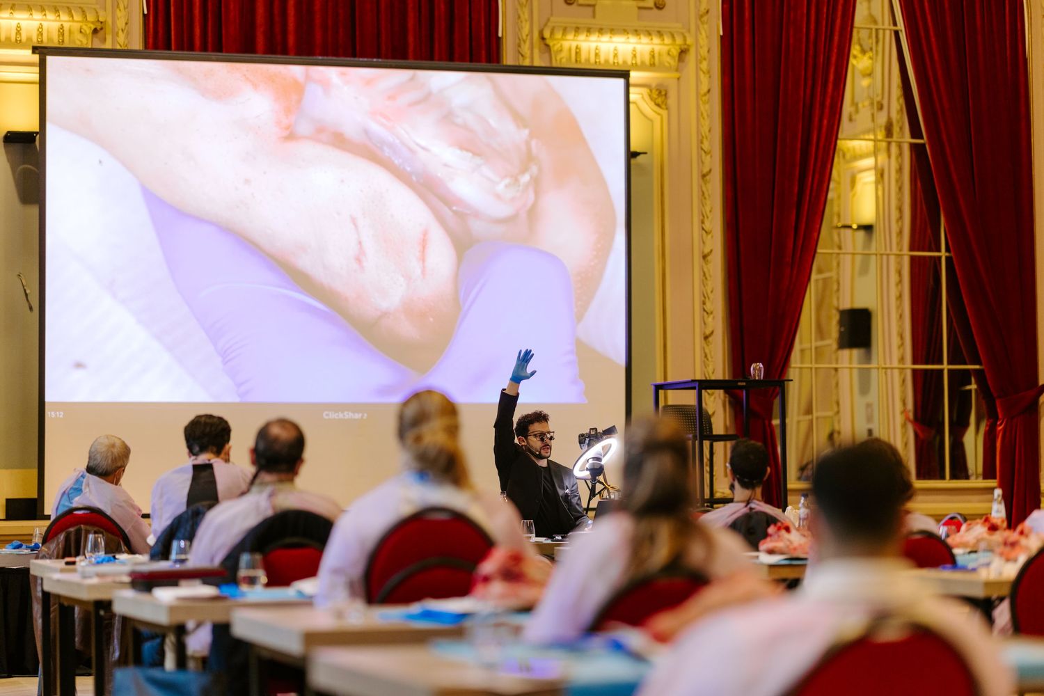 Medical professional presents surgical procedure on large screen to seated attendees in ornate conference room.