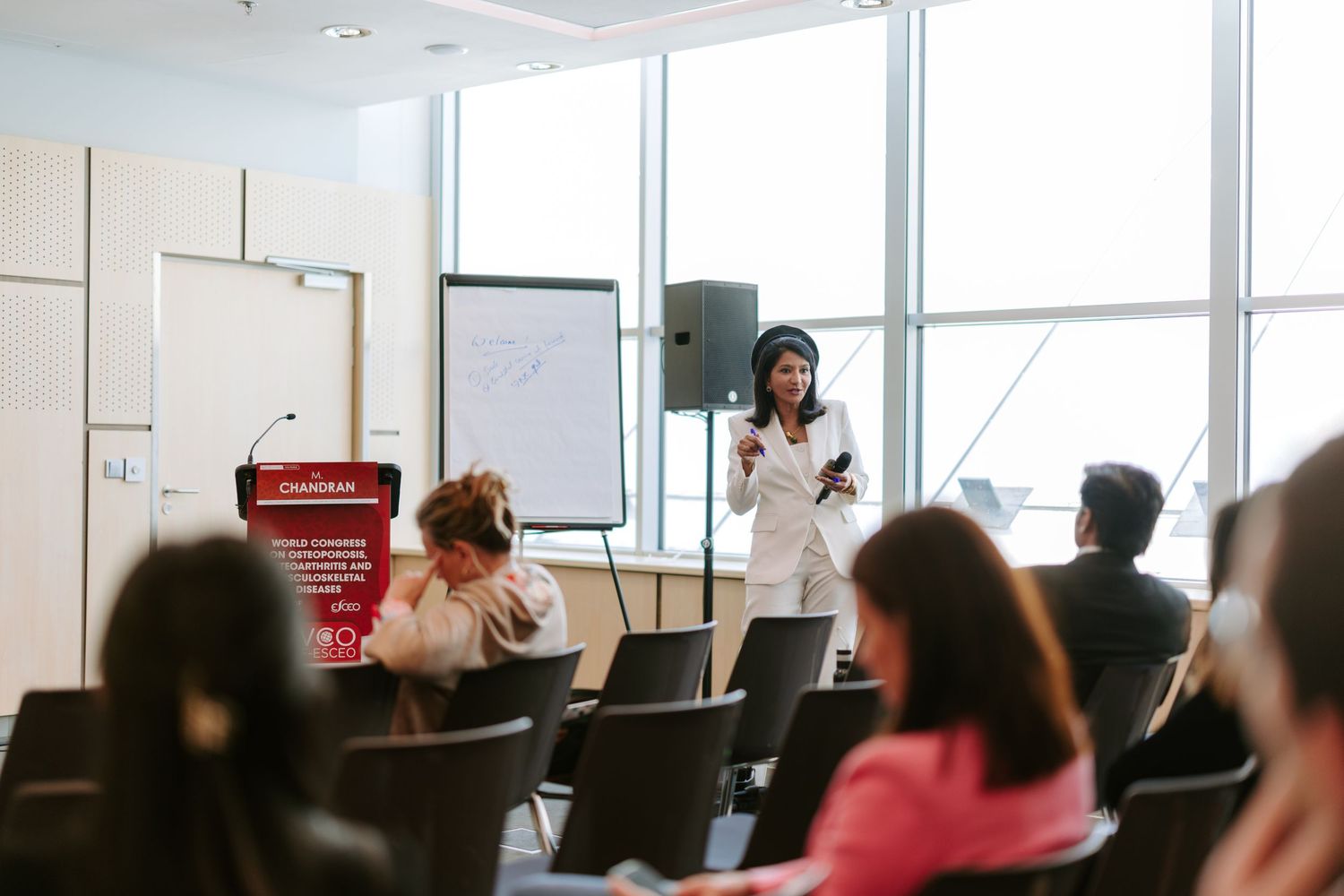 Female speaker presenting at a business conference to seated attendees in a modern meeting room.