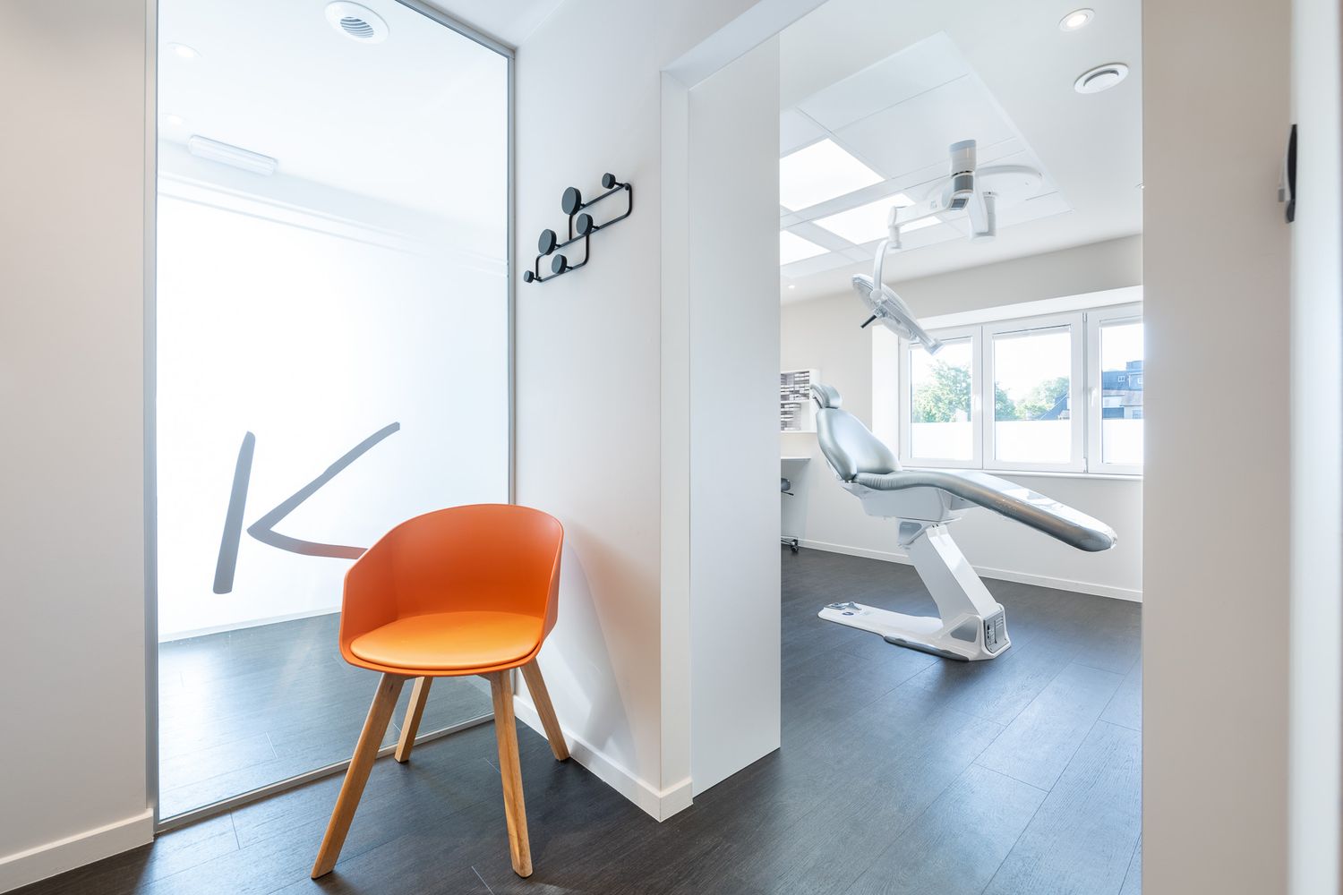 Modern dental office entrance with an orange chair, frosted glass K logo door, and a dental chair visible inside.