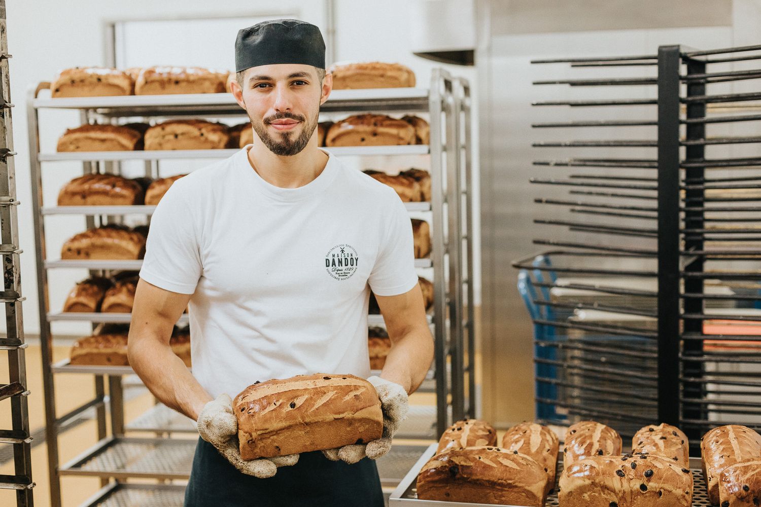 Young male baker in white t-shirt holding freshly baked bread loaves in a professional bakery.