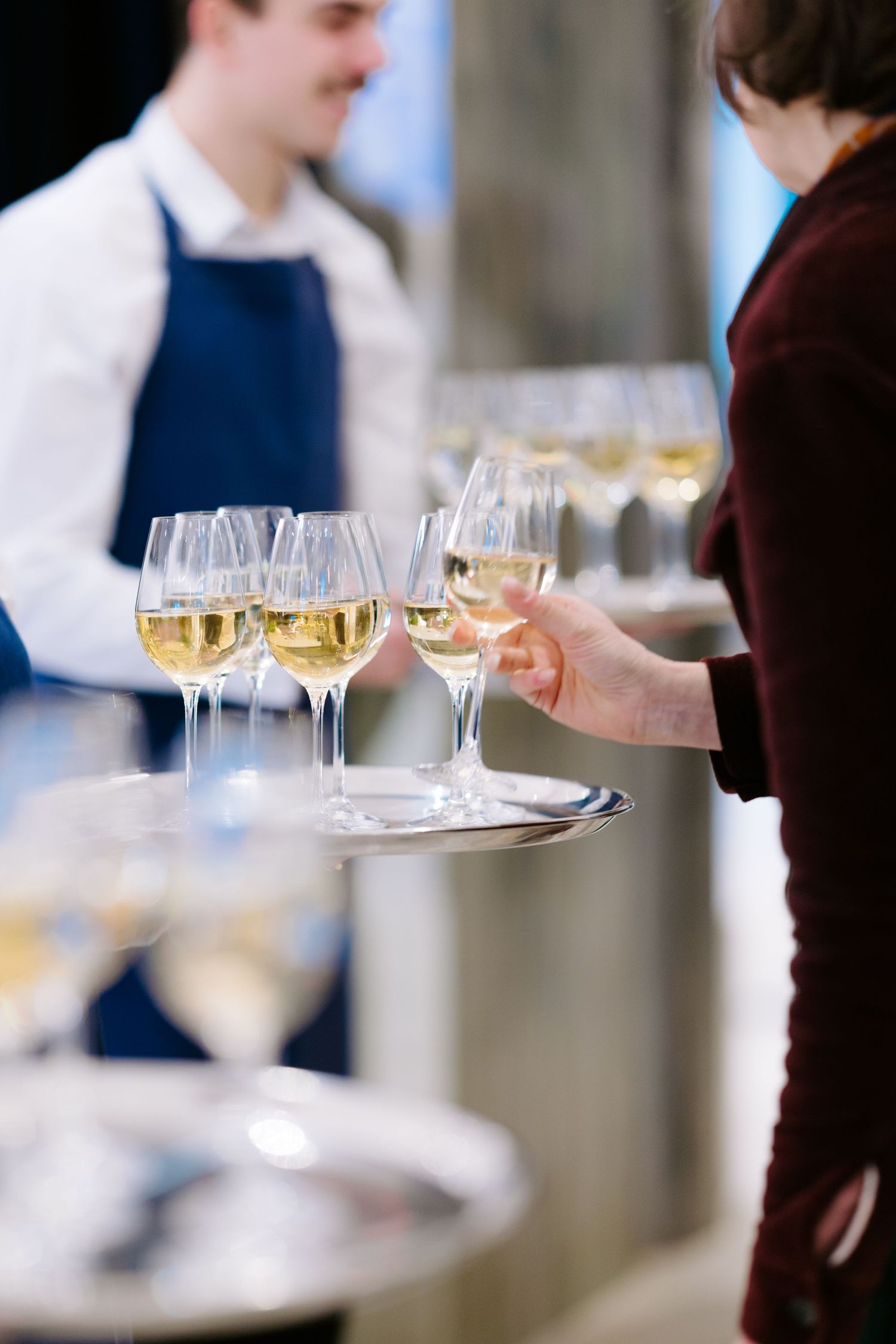 Waiter in blue apron serving white wine glasses on silver tray at elegant catered event.