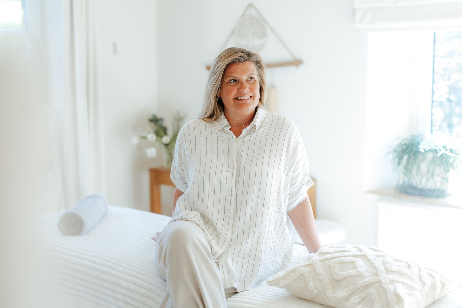 Smiling middle-aged woman sitting on a white bed in a bright, minimalist bedroom.