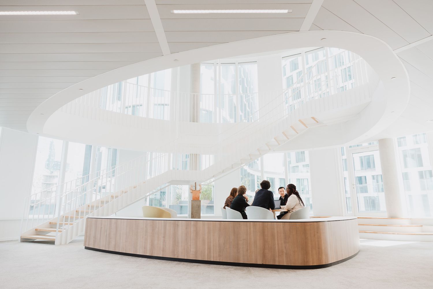 Modern bright white building interior with a curved staircase and people gathered at a wooden reception desk.