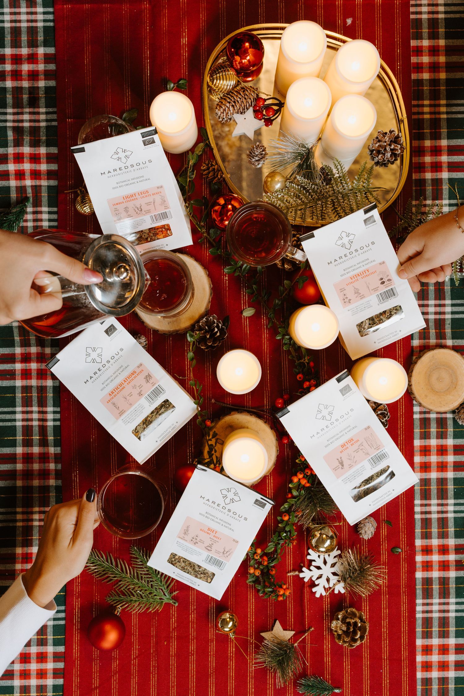 Top view of a festive Christmas table with tea packets, candles, ornaments, and hands holding glasses of tea.