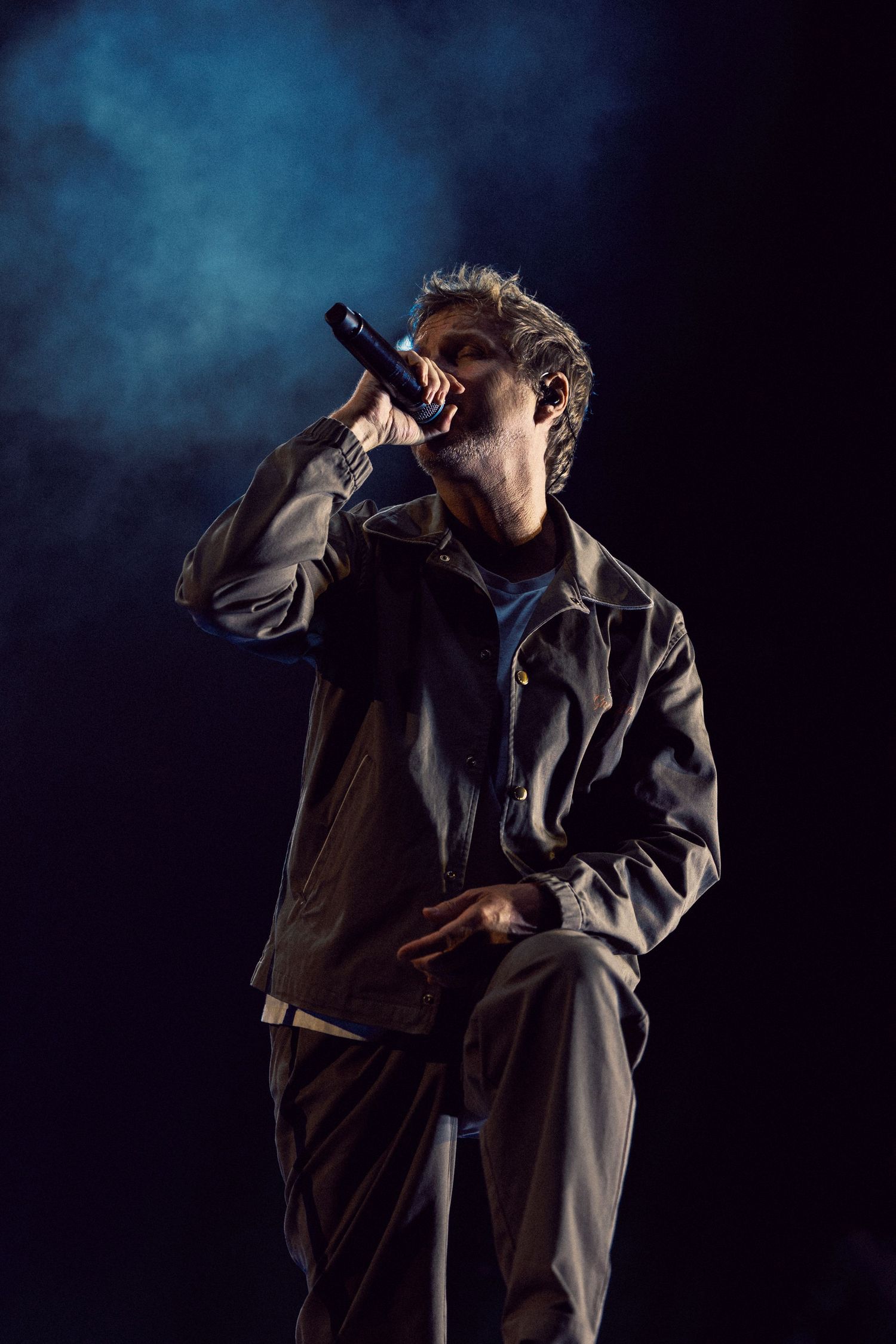 Male rapper performing on dark stage with microphone, wearing gray jacket, dramatic blue smoke lighting in background.
