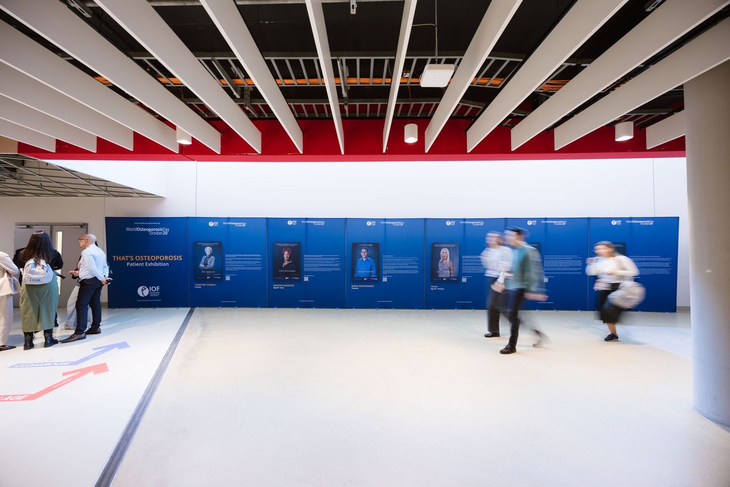 Visitors walk past a blue exhibition display wall in a bright modern conference hall.