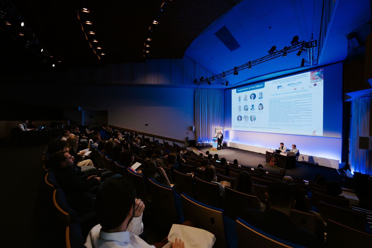 Large conference auditorium with audience attending a professional presentation with a bright projected screen on stage.