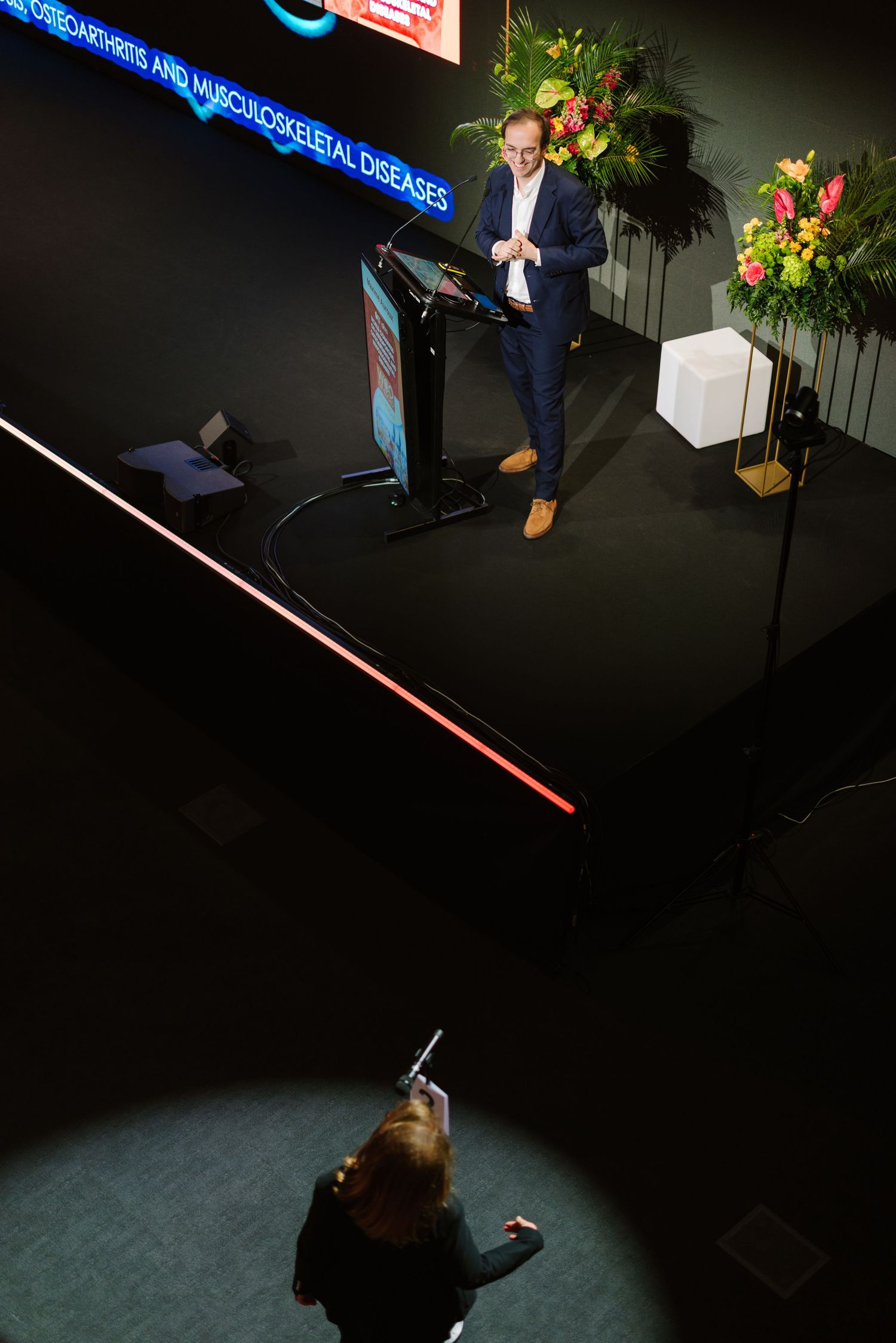 Speaker presents at osteoarthritis conference podium on dark stage with floral arrangements, viewed from above.