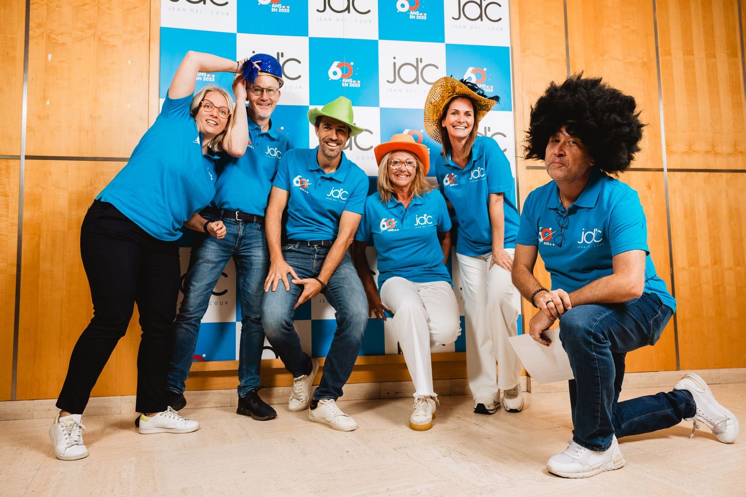 Energetic group of six people in matching blue JDC shirts posing playfully in front of a branded backdrop.