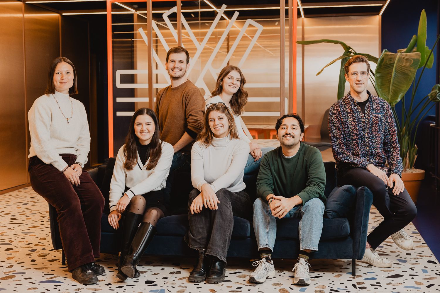 Group of seven young professionals smiling together on a couch in a modern office.