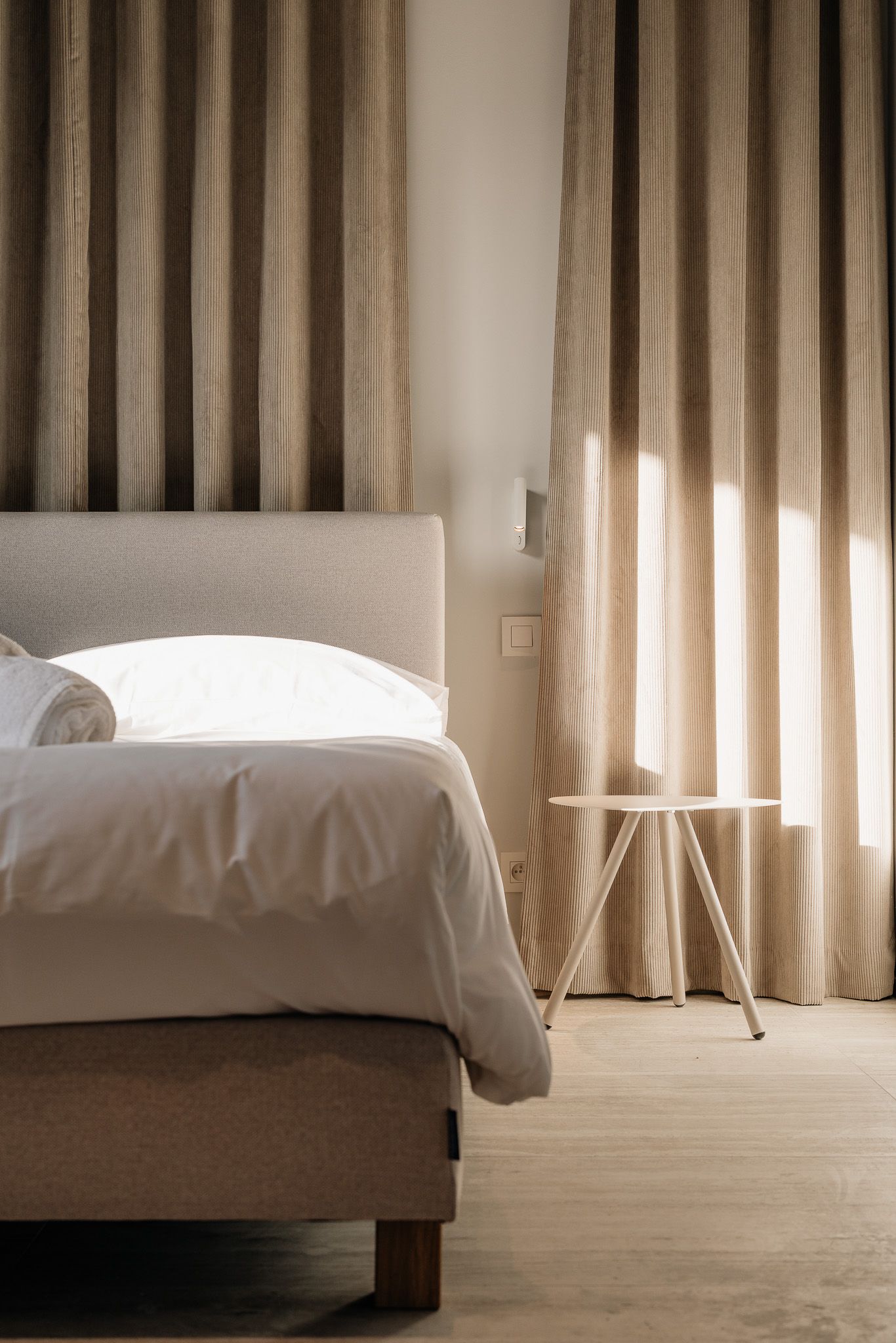 Serene hotel bedroom with upholstered headboard, white linen, small side table, and warm sunlit striped curtains.