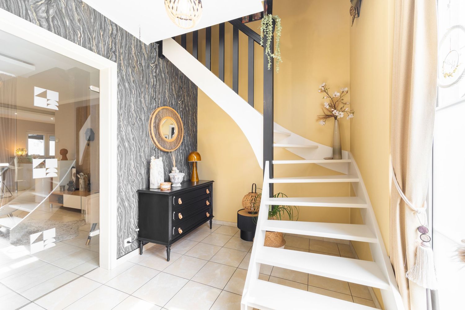 Bright home entryway featuring white staircase, black dresser, decorative mirror, and textured wallpaper accent wall.