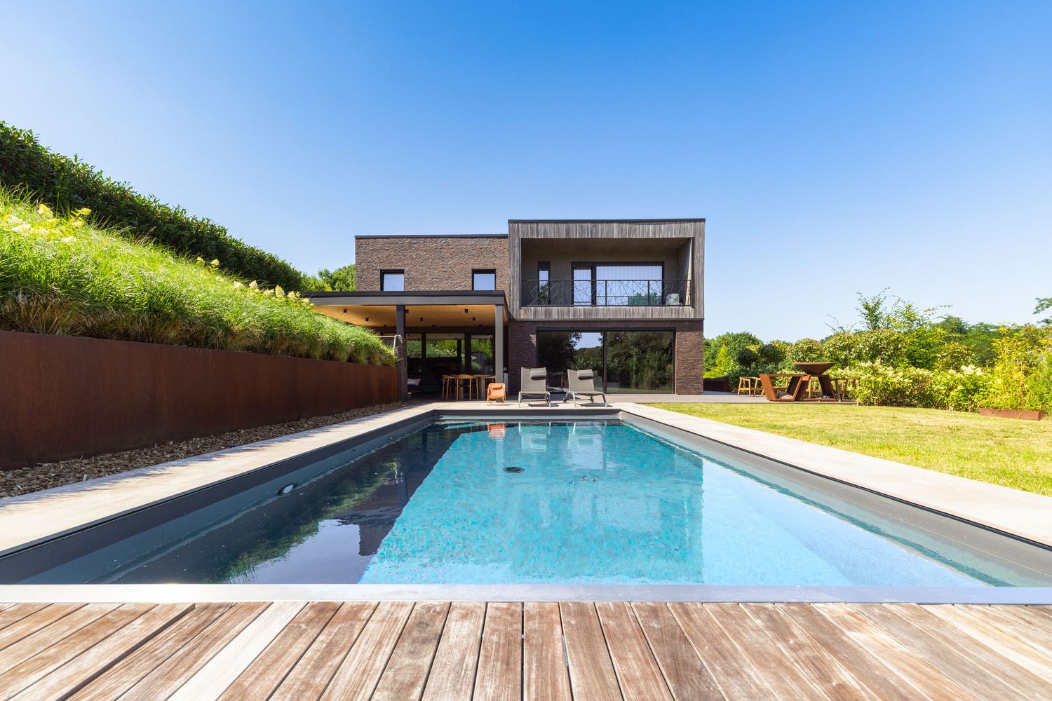 Modern two-story home with sleek pool and wooden deck surrounded by lush greenery under a clear blue sky.