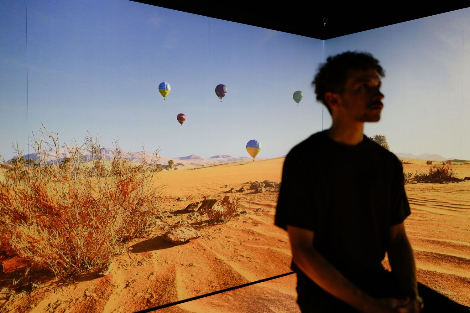 A young man standing in silhouette before a large desert landscape projection with hot air balloons.