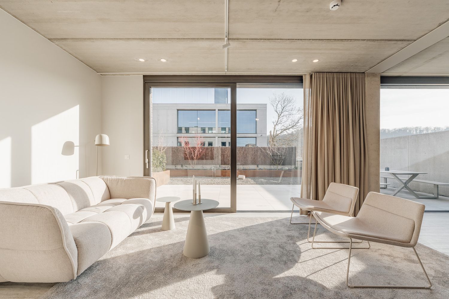 Bright minimalist living room with white furniture and large sliding doors to a sunny terrace.