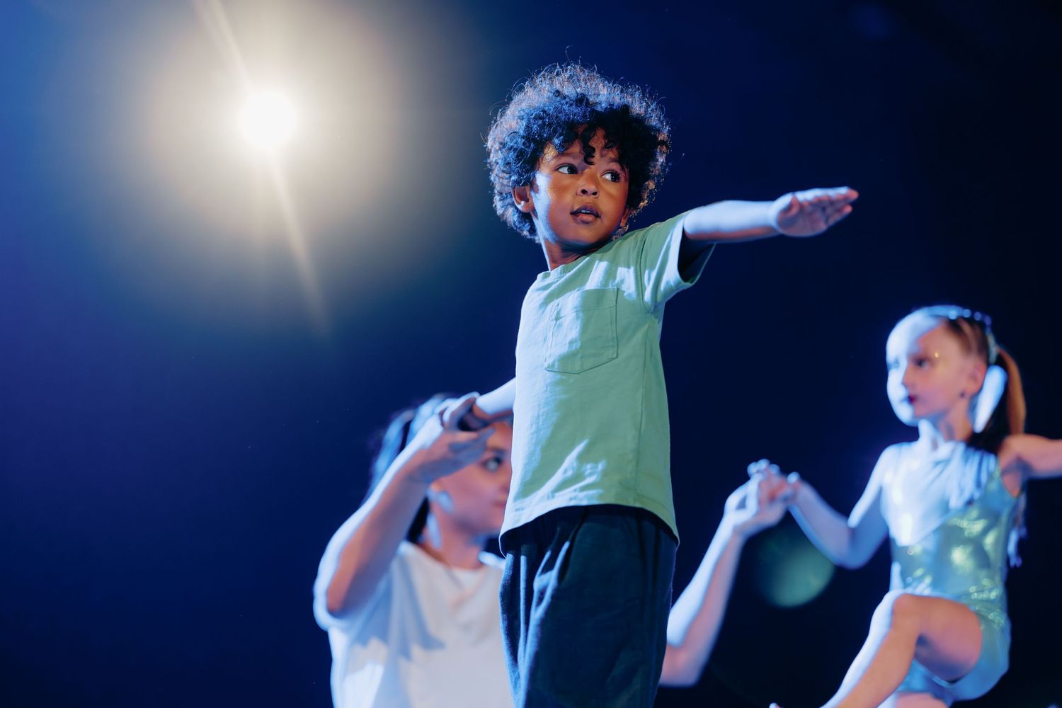 Young curly-haired boy dancing on stage under spotlight with other children performers in background.