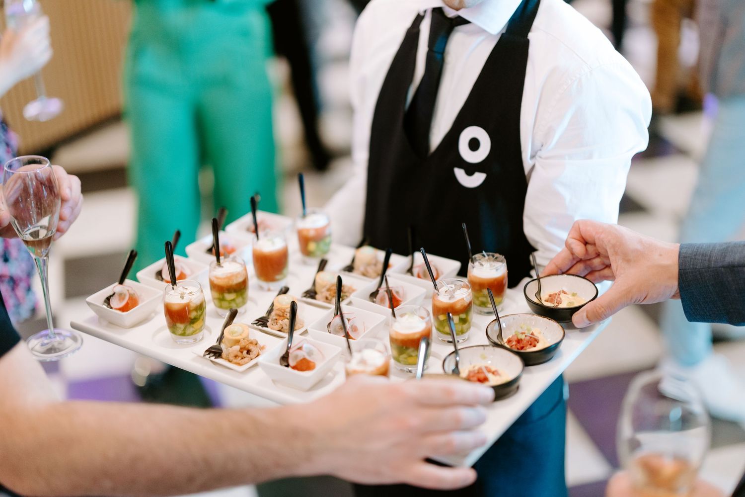 Catering server holding tray of elegant appetizers in small glasses at a formal event or reception.