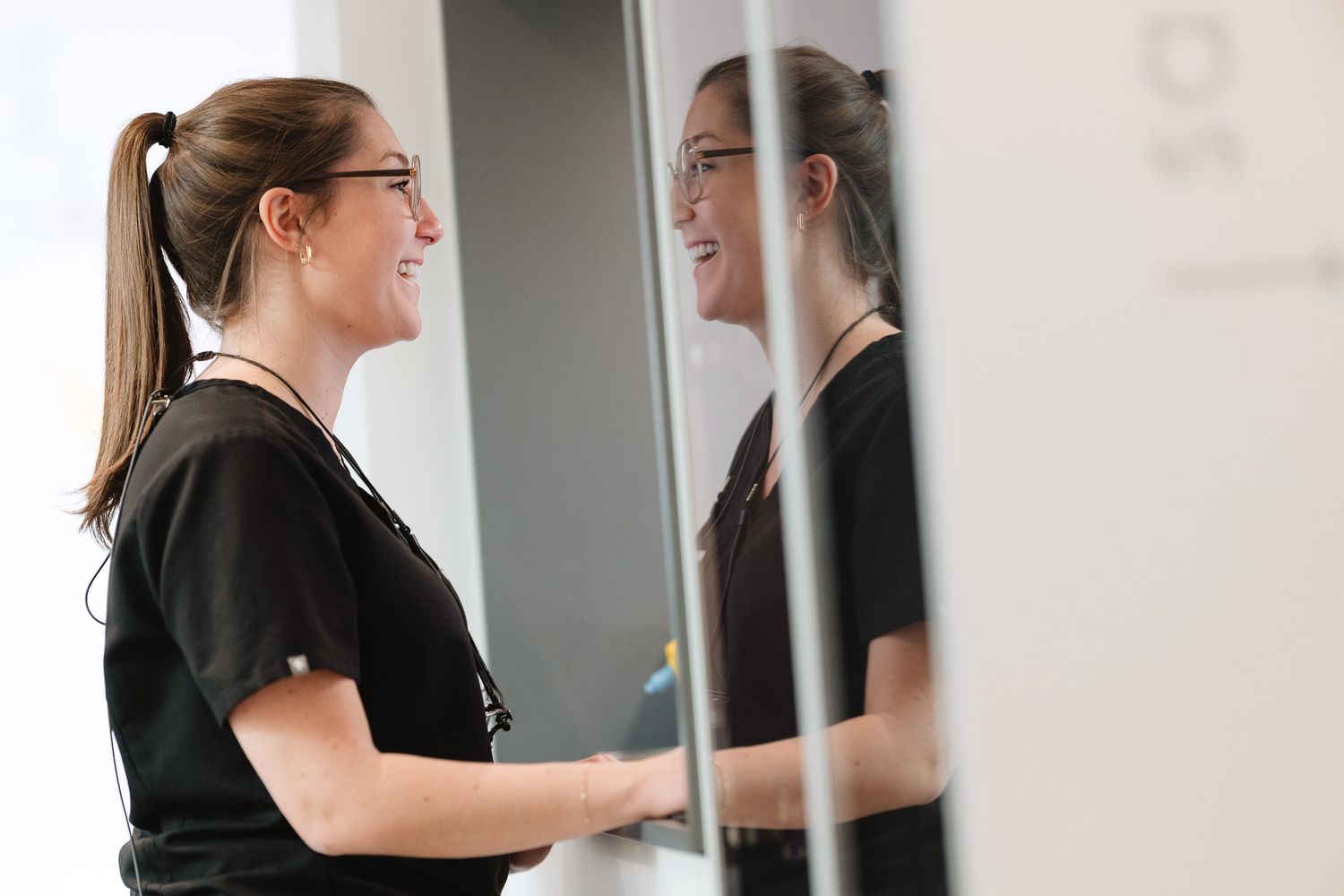 Smiling female healthcare professional in black scrubs reflected in glass partition in clinic.