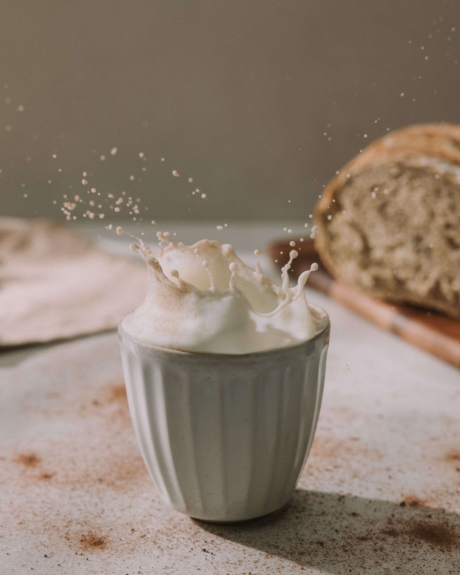 Milk splashing out of a white ceramic cup with bread loaf in the background.