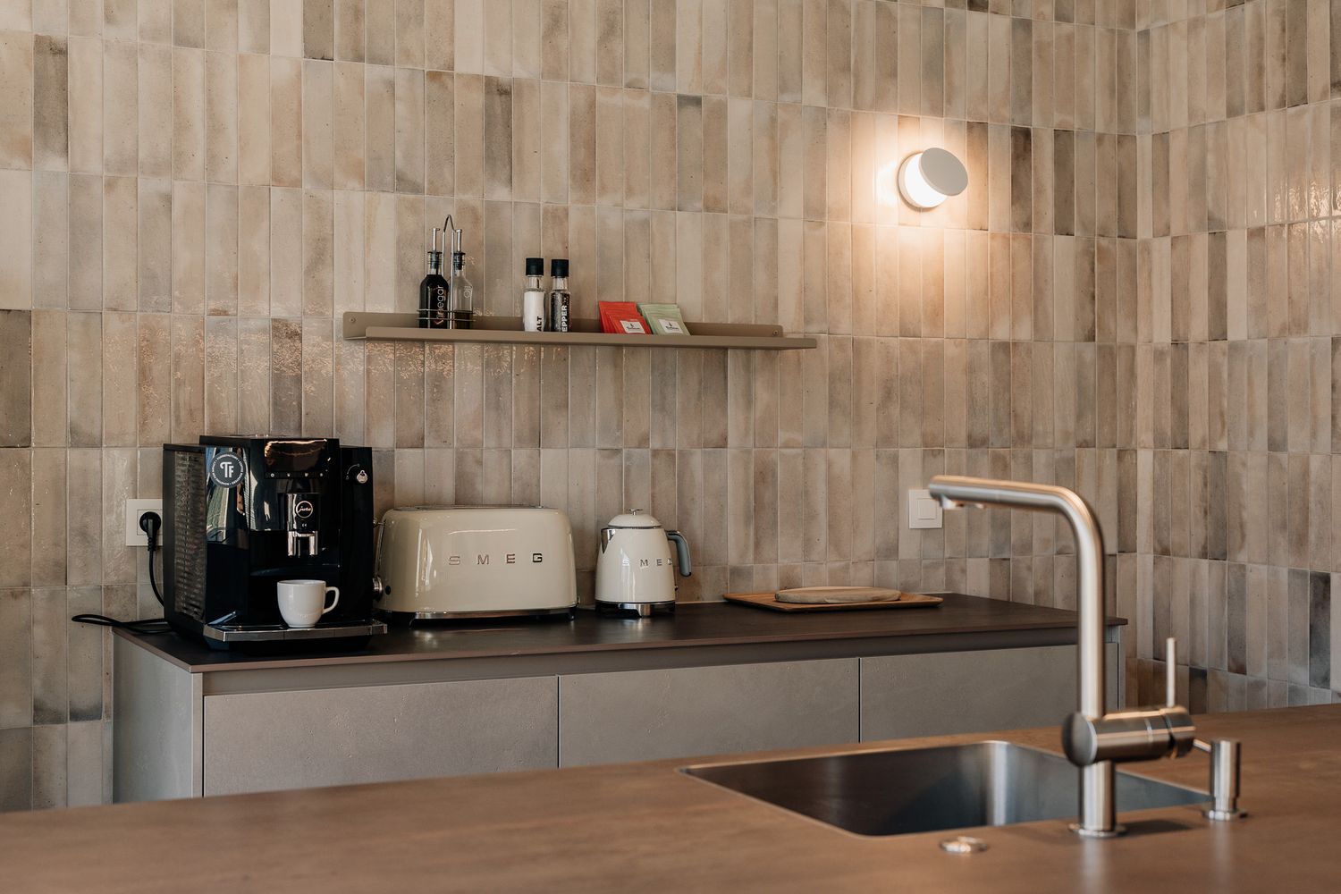 Contemporary kitchen counter with coffee machine, sink, and wooden tile backsplash illuminated by a wall-mounted light.