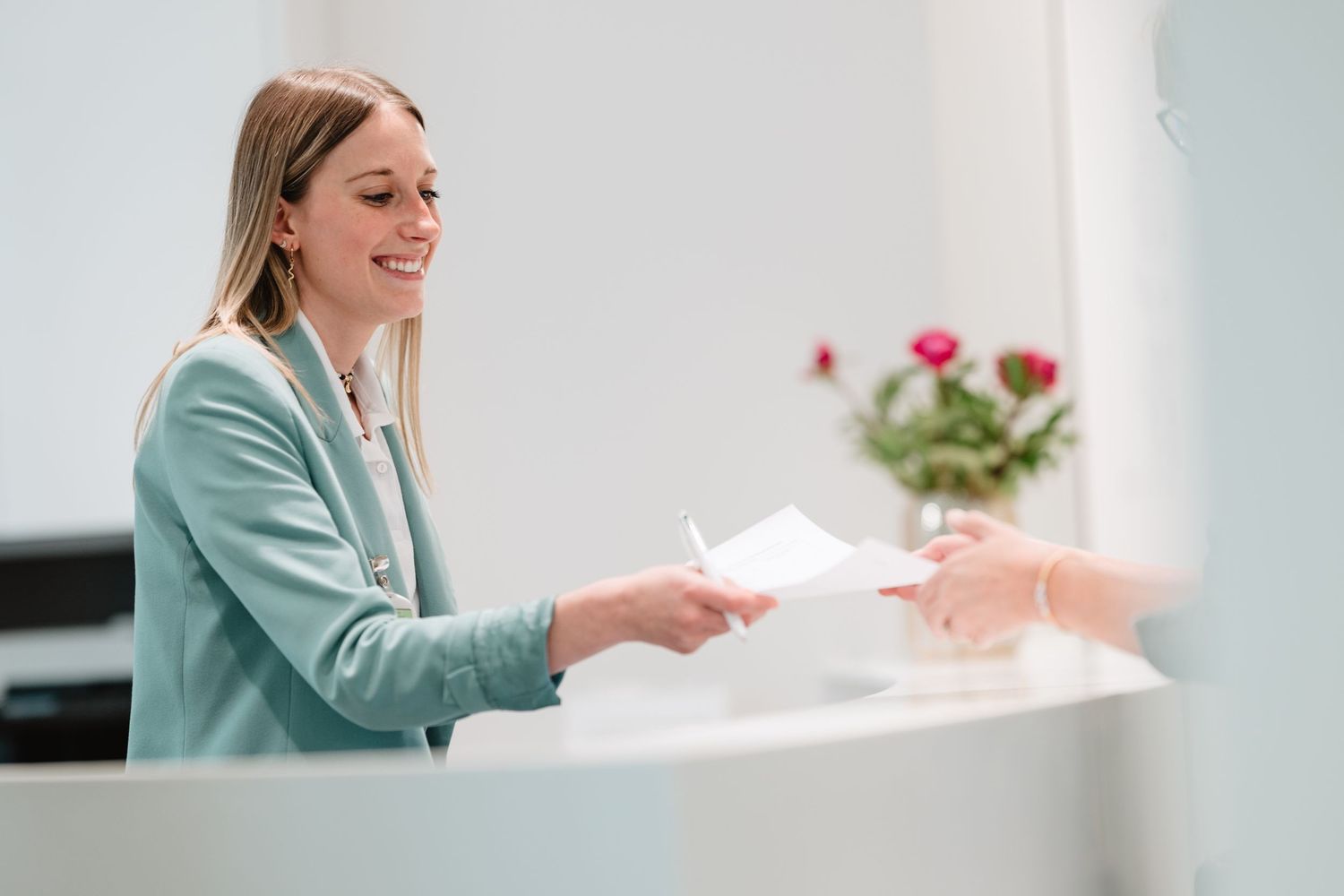 Smiling blonde receptionist handing documents to a client at a bright modern reception desk with pink flowers.