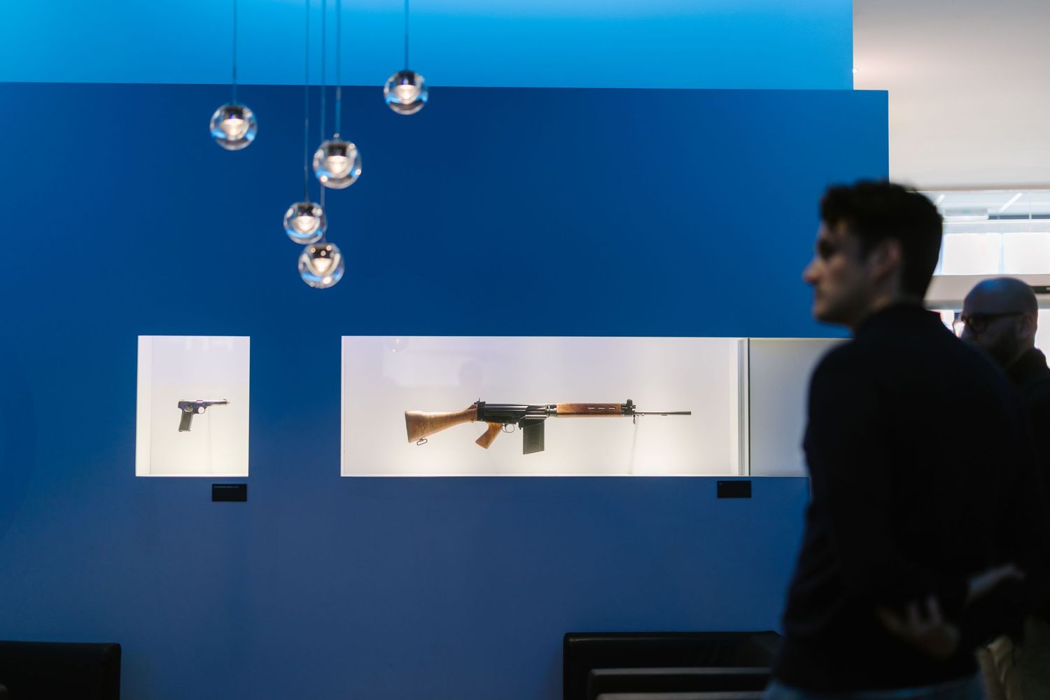 Rifle displayed in illuminated case at firearms exhibition with decorative pendant lights.