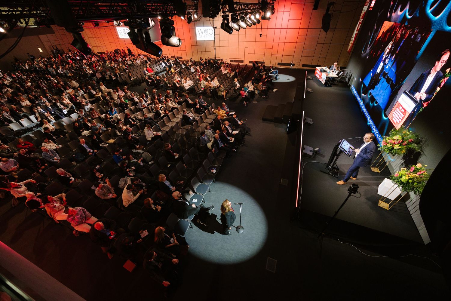 Speaker presents on stage to a large packed audience at a professional conference event.