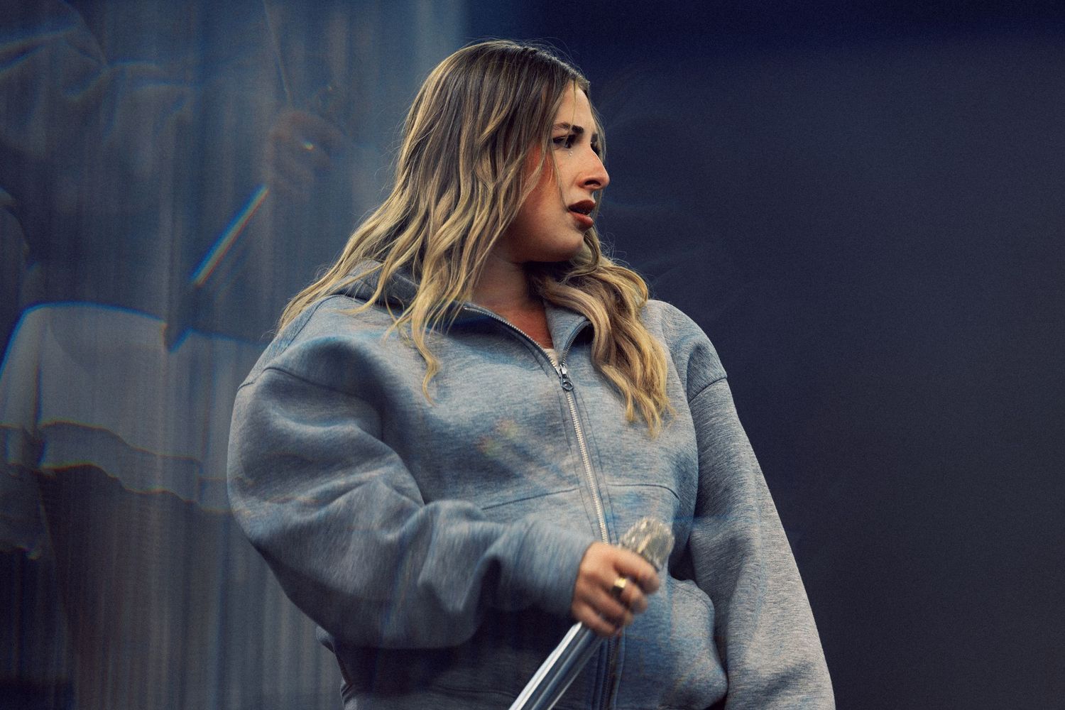 Young woman with blonde hair wearing an oversized gray zip-up fleece jacket, holding a microphone stand on stage.