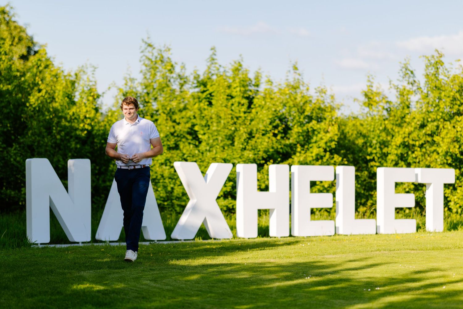 Person posing in front of large white decorative NAXHELET letters on a green golf course lawn.