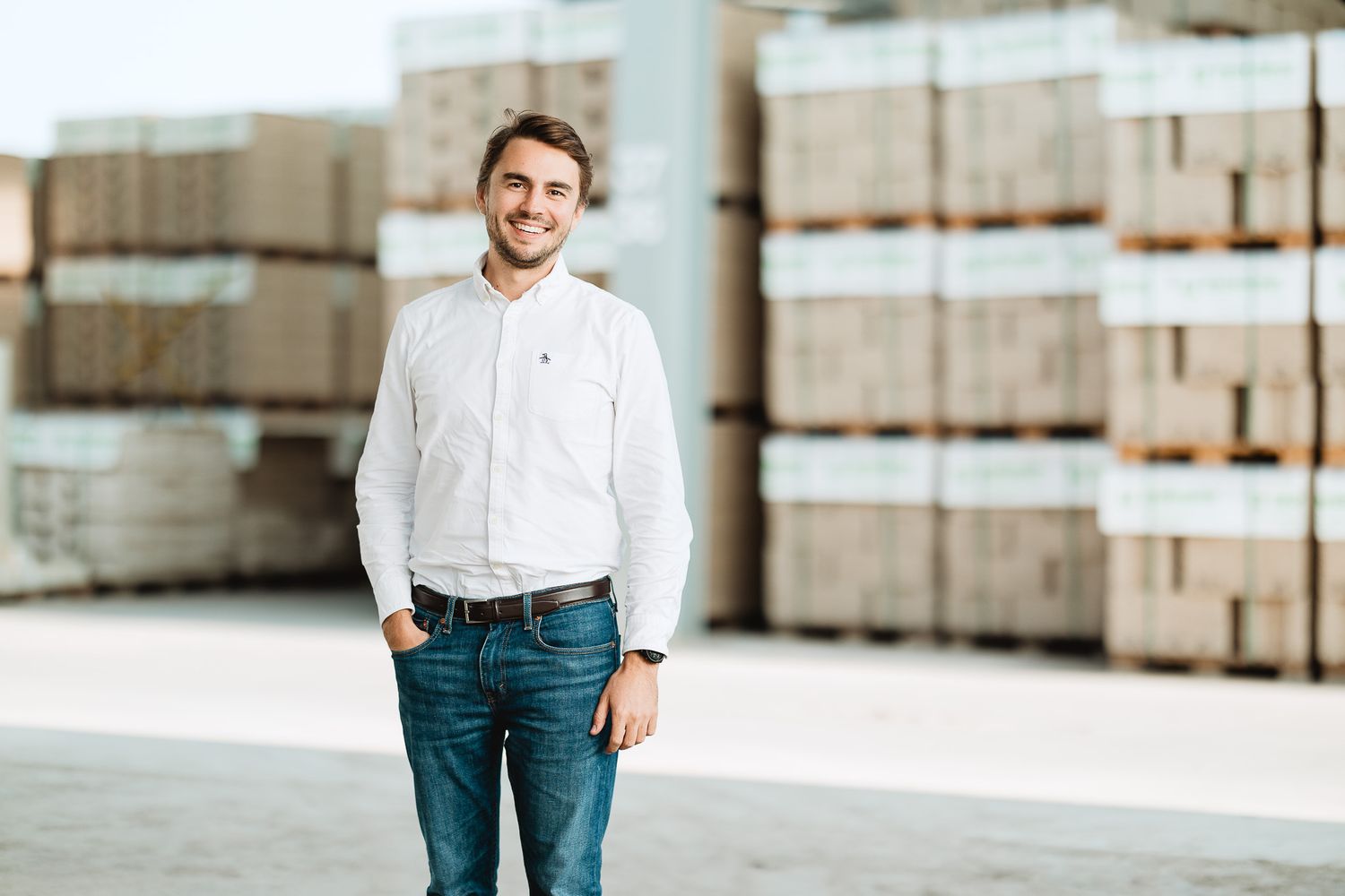 Smiling young businessman in white shirt standing in front of stacked pallets in a warehouse.