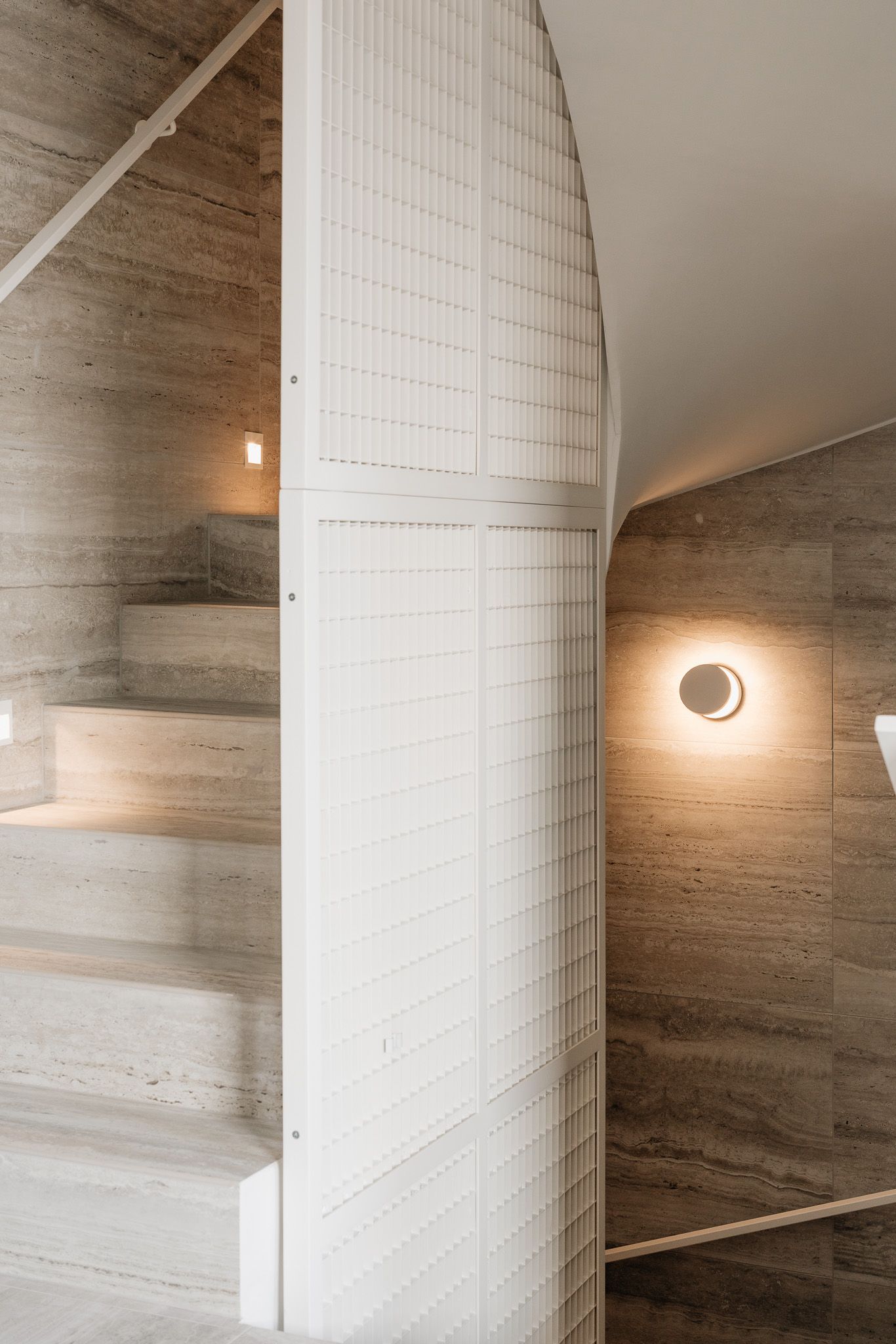 Elegant staircase with arched white tiled column, travertine stone walls, and warm sconce lighting in modern interior.